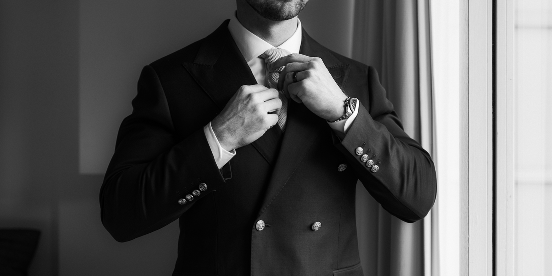 Black-and-white photograph of a man adjusting his tie while wearing a forrest green double-breasted suit beside a window.