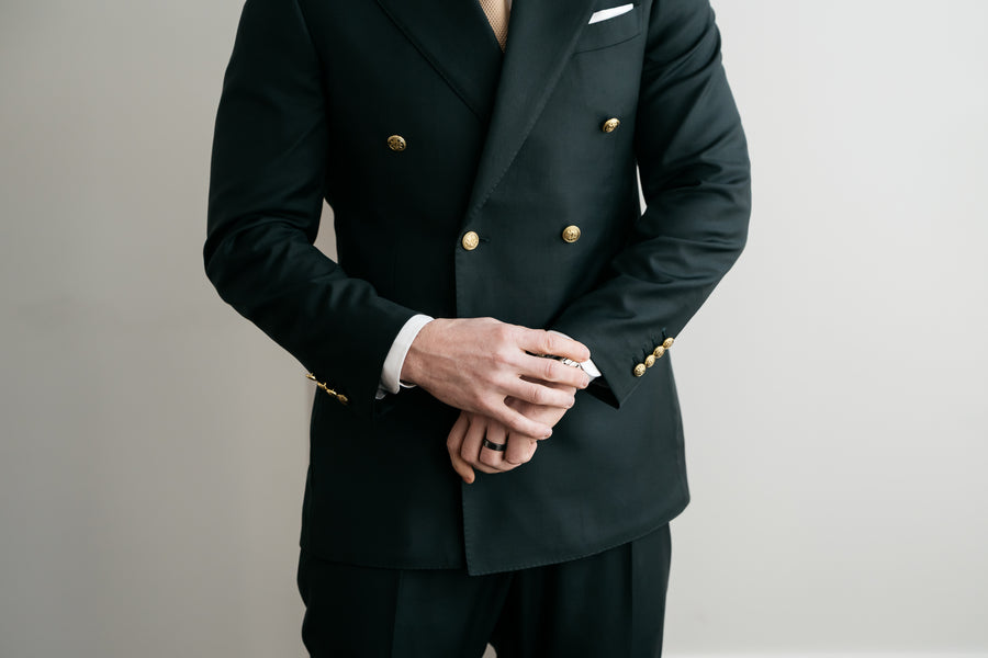 A close-up of a person wearing a forrest green double-breasted suit jacket with gold buttons, a white pocket square, and matching trousers, standing against a light gray background.