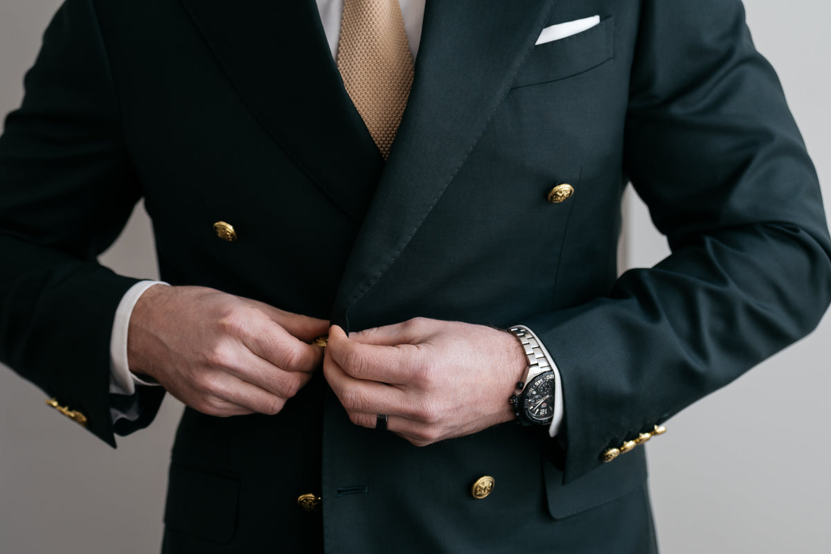 A close-up of a man's hands buttoning a dark green double-breasted suit jacket with gold buttons, paired with a beige tie and white pocket square, set against a neutral gray background.