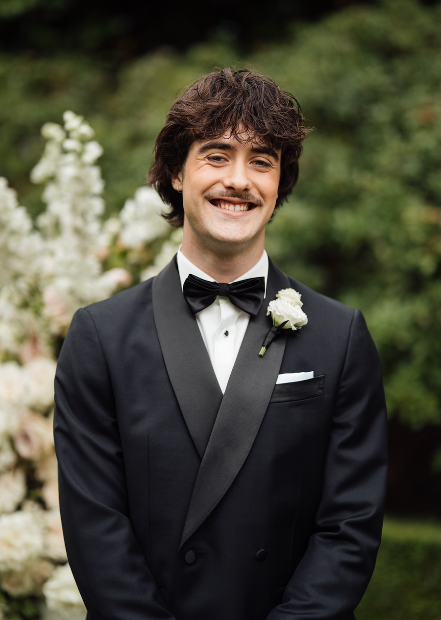 A smiling man wearing a black double-breasted tuxedo with black satin lapels, a black bow tie, a white dress shirt, and a white boutonniere, photographed outdoors against a soft-focus green garden background.