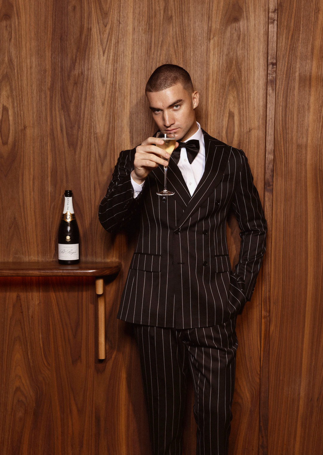 Man wearing a black double-breasted pinstripe suit with a white shirt and black bow tie, holding a champagne glass.