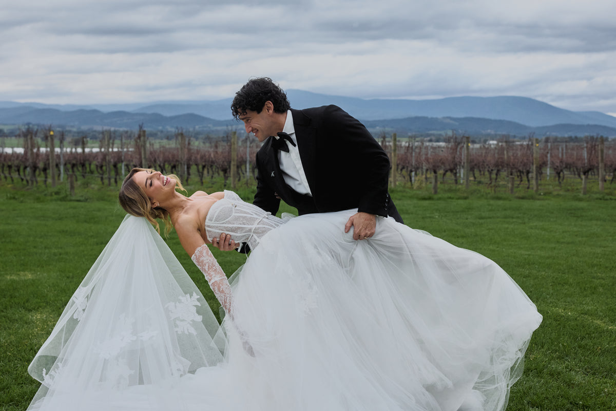 A bride in a white lace wedding gown and veil is being lifted by a groom in a black tuxedo while standing in a vineyard with mountains in the background.