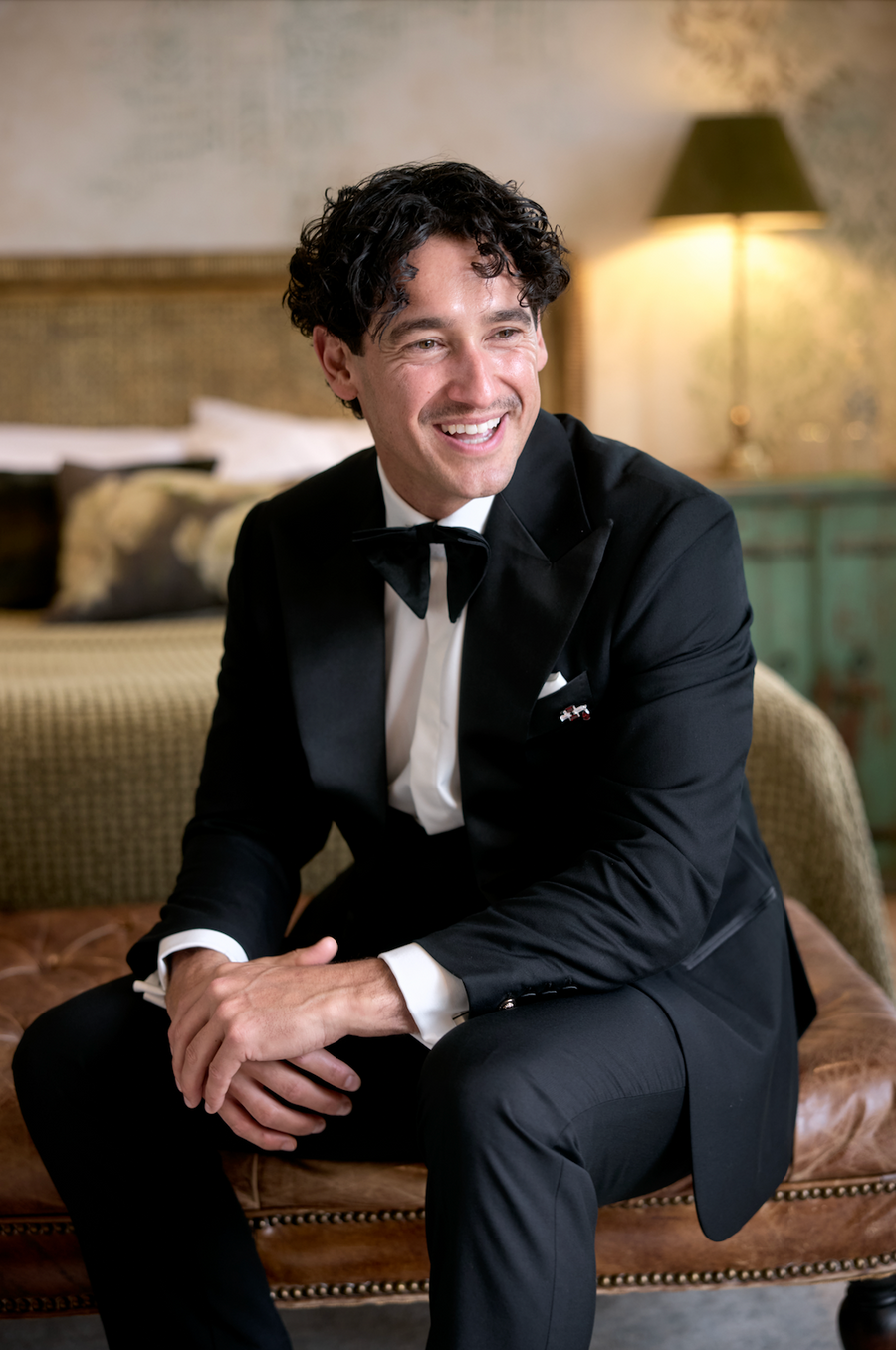 A man wearing a black tuxedo with a white shirt and black bow tie, sitting on a brown leather bench in a warmly lit bedroom setting.