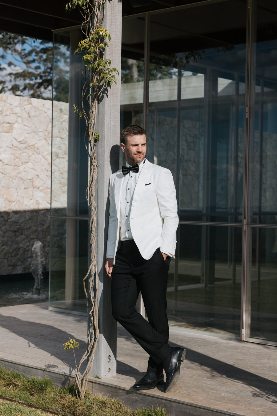A man wearing a white tuxedo jacket with black trousers and a black bow tie, leaning against a modern glass-walled building next to a climbing vine.